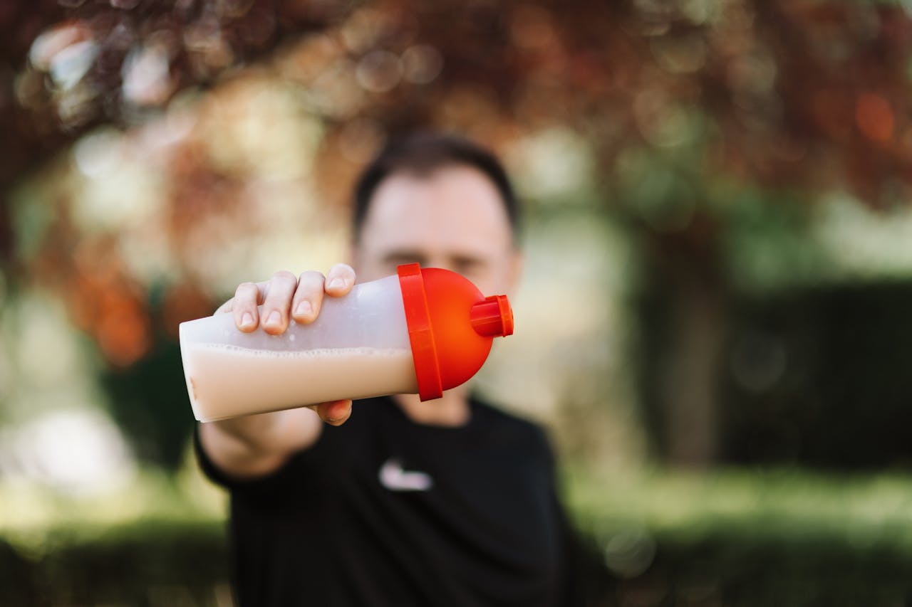 Focus on a man holding a plastic protein shake tumbler outdoors with a blurred background.